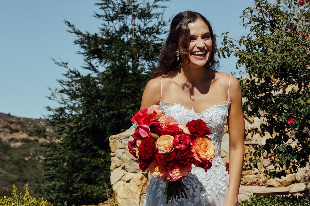 Smiling bride holding vibrant red and orange rose bouquet in floral wedding dress outdoors on sunny day.