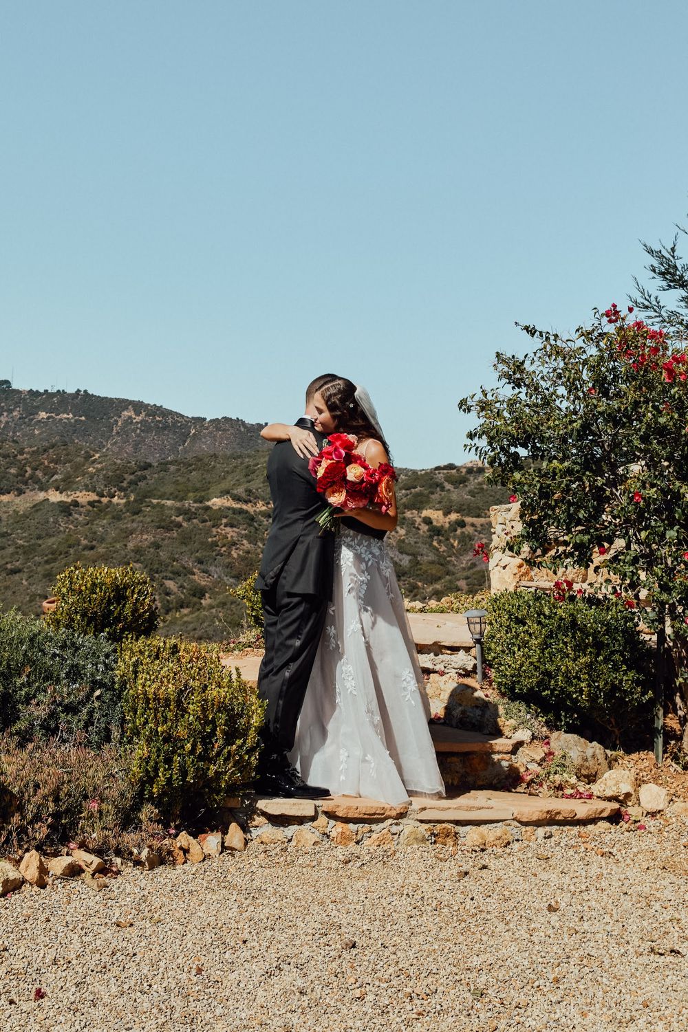 A bride and groom embrace outdoors with red bouquet, mountain backdrop, and blue sky at a rustic wedding venue.