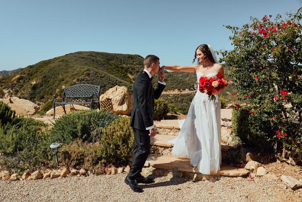 Bride in white gown holds red bouquet reaching toward groom in black suit on scenic hillside path.