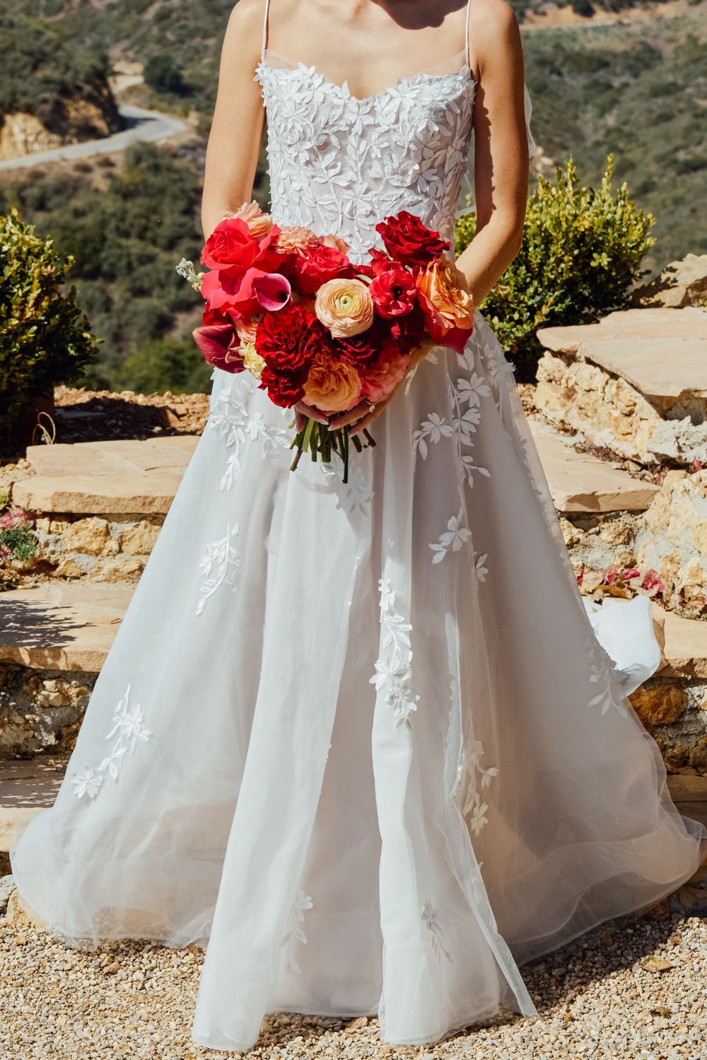 Bride in a white lace A-line wedding gown holding a vibrant red and orange bouquet outdoors.