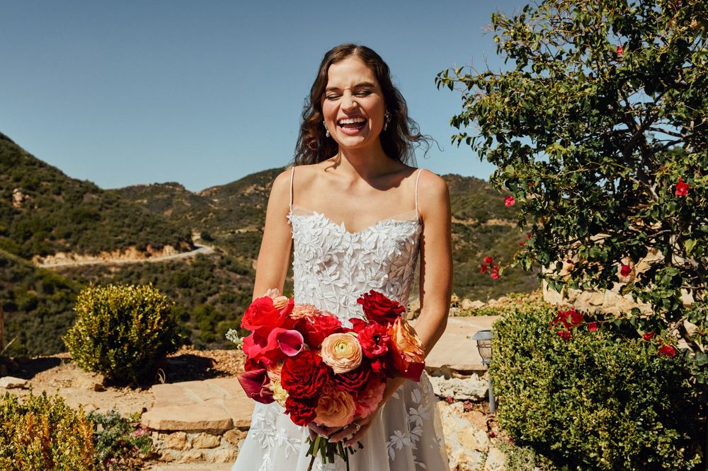 Smiling bride in strapless lace gown holding vibrant red and orange bouquet outdoors with mountain backdrop.