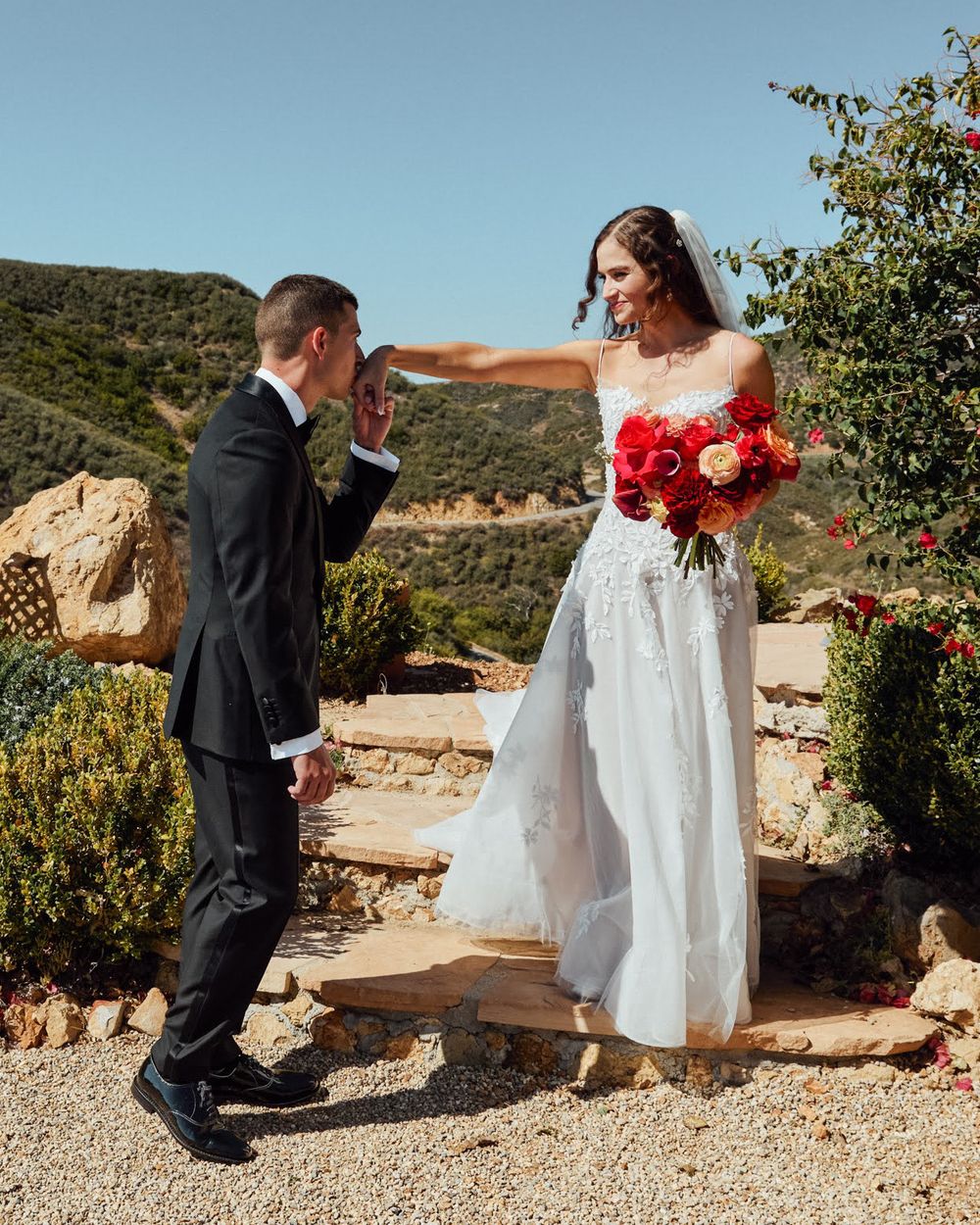 Groom kissing bride's hand in white off-shoulder gown with red bouquet at outdoor wedding ceremony.