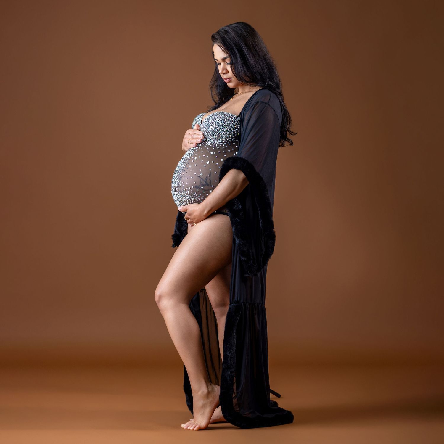 Pregnant woman in a beaded bodysuit and black sheer robe poses gracefully against a warm brown studio background.