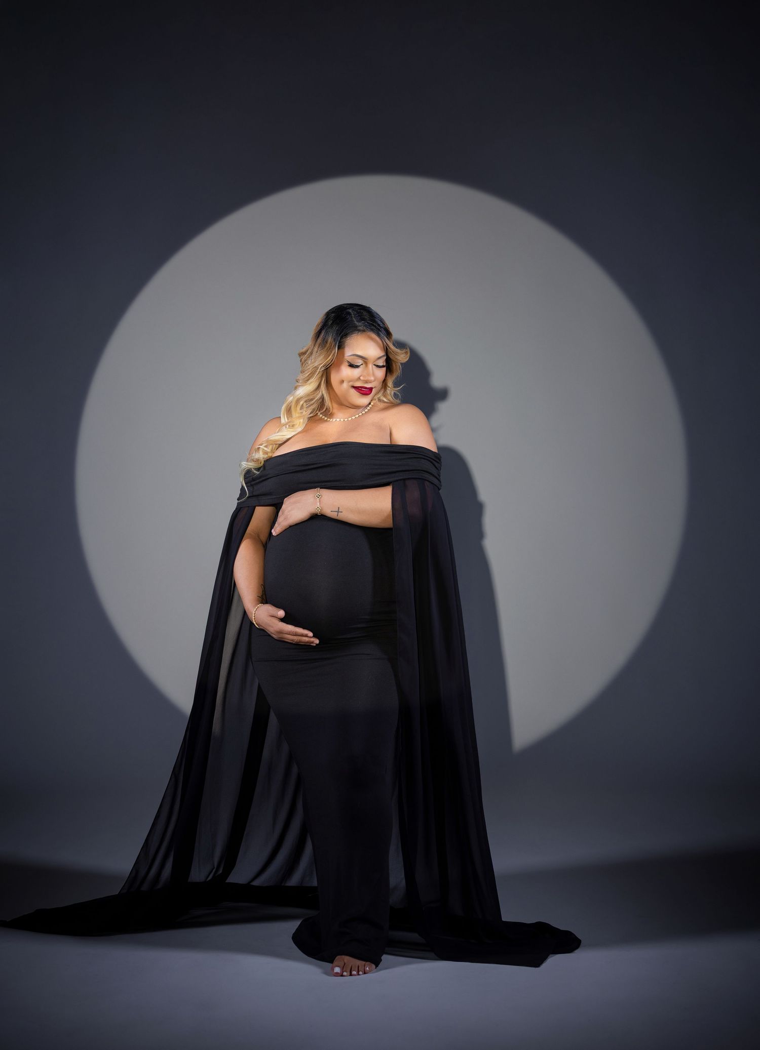 Pregnant woman in elegant black off-shoulder gown with cape, posing in studio with circular spotlight backdrop.