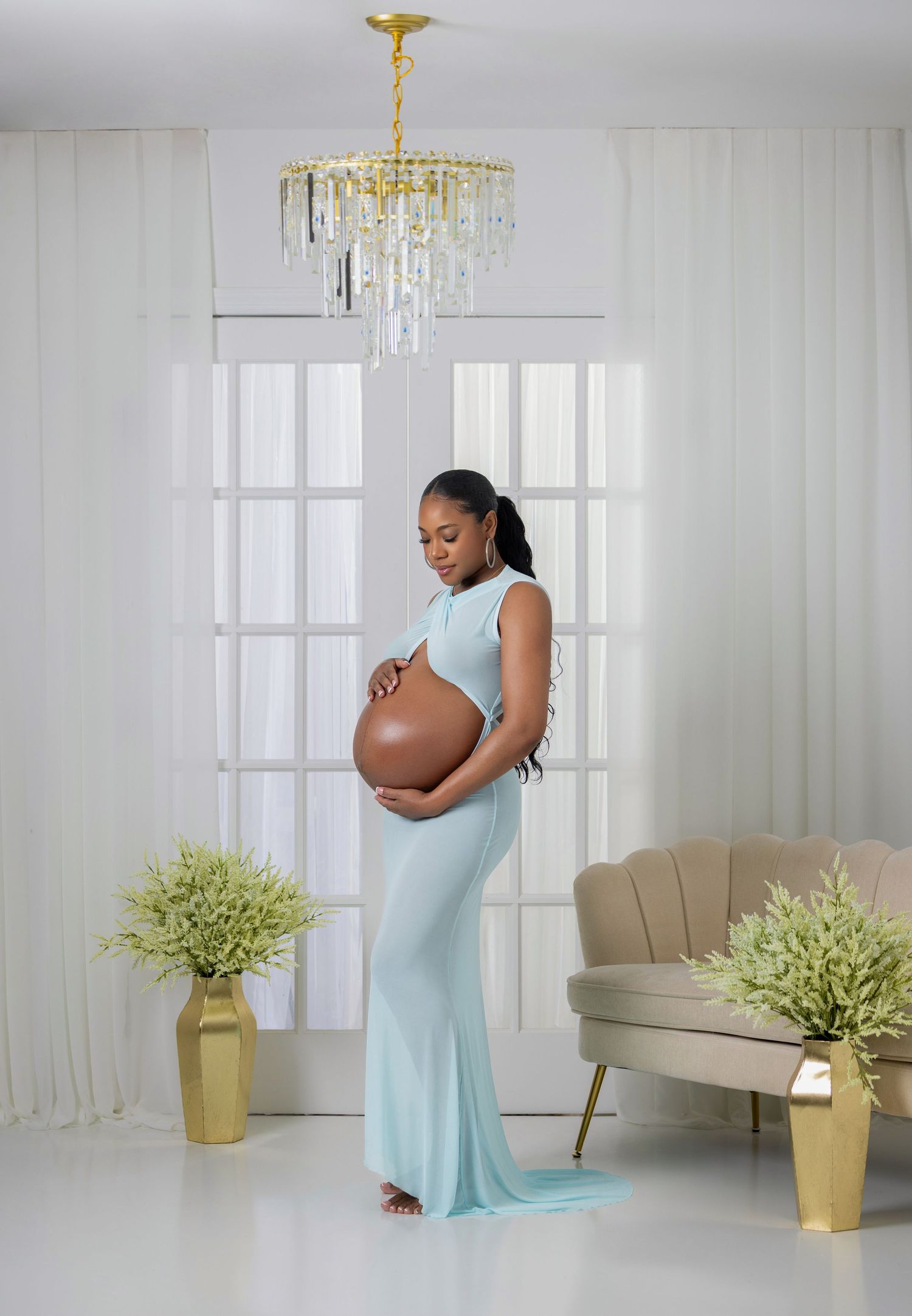 Pregnant woman in light blue maternity gown cradling baby bump in elegant white studio with chandelier.