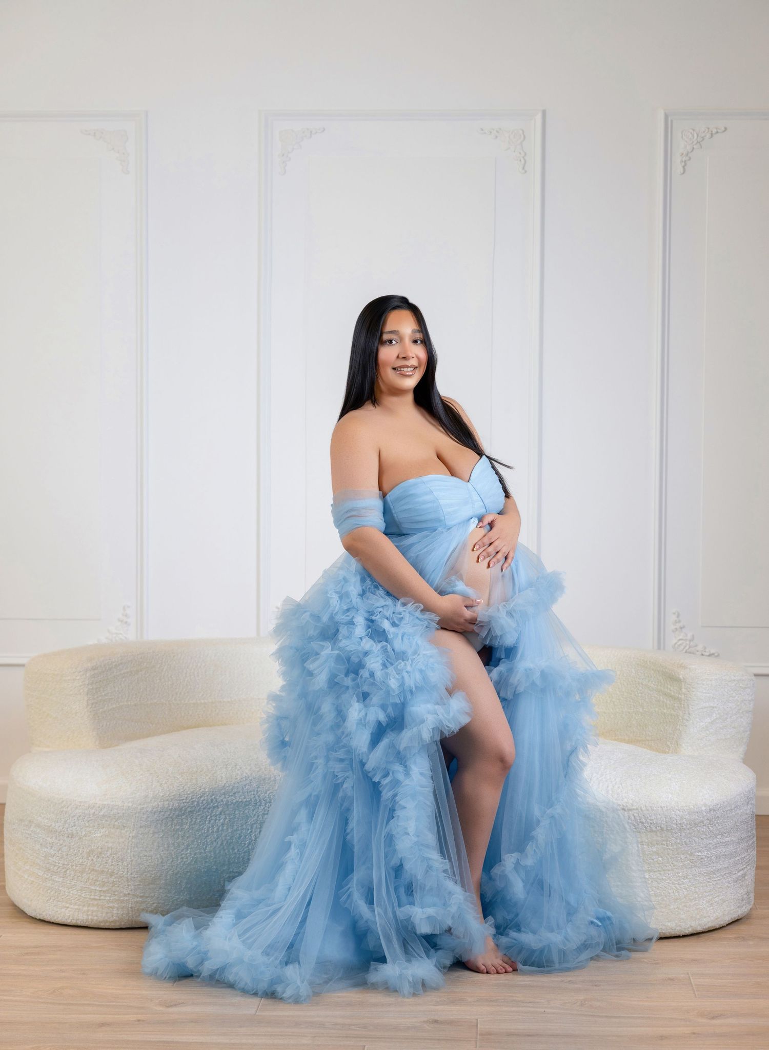 Pregnant woman in a flowing blue tulle off-shoulder gown poses gracefully in a bright white studio setting.
