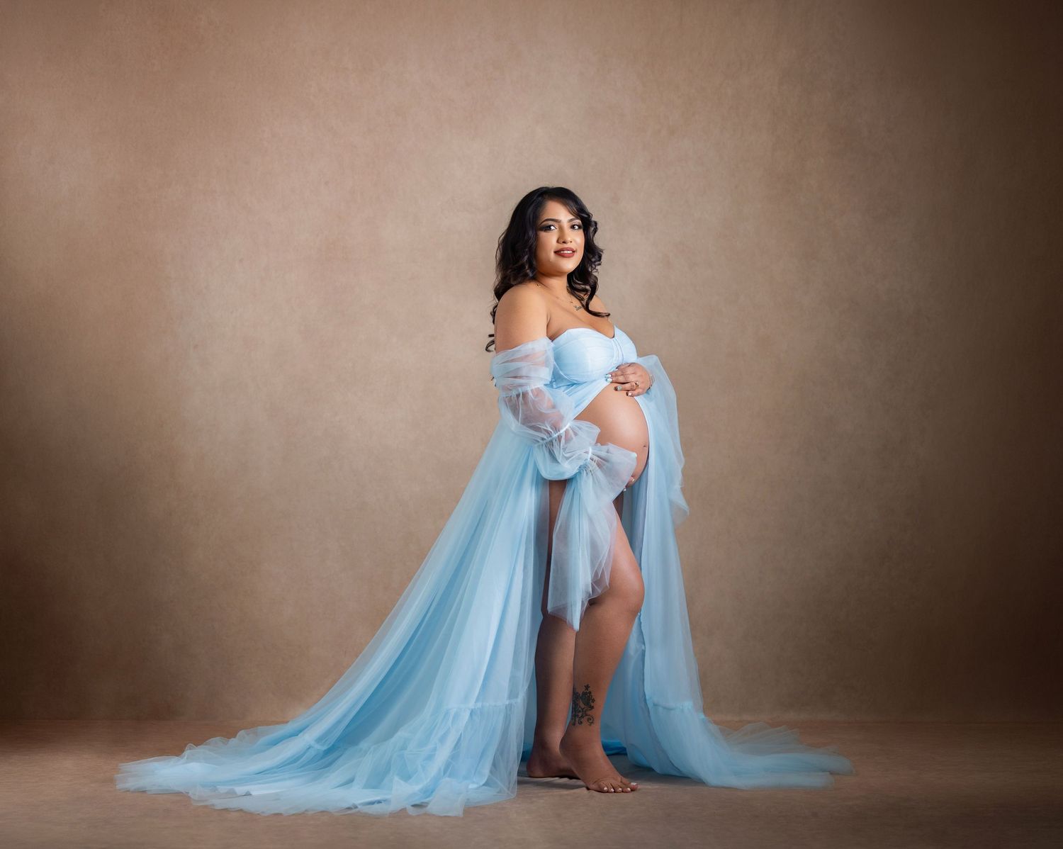 Pregnant woman in elegant light blue tulle maternity gown posing gracefully against a warm beige studio backdrop.