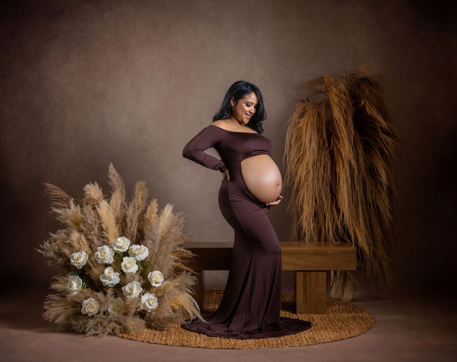 Pregnant woman in brown off-shoulder gown poses in studio with pampas grass and white roses.