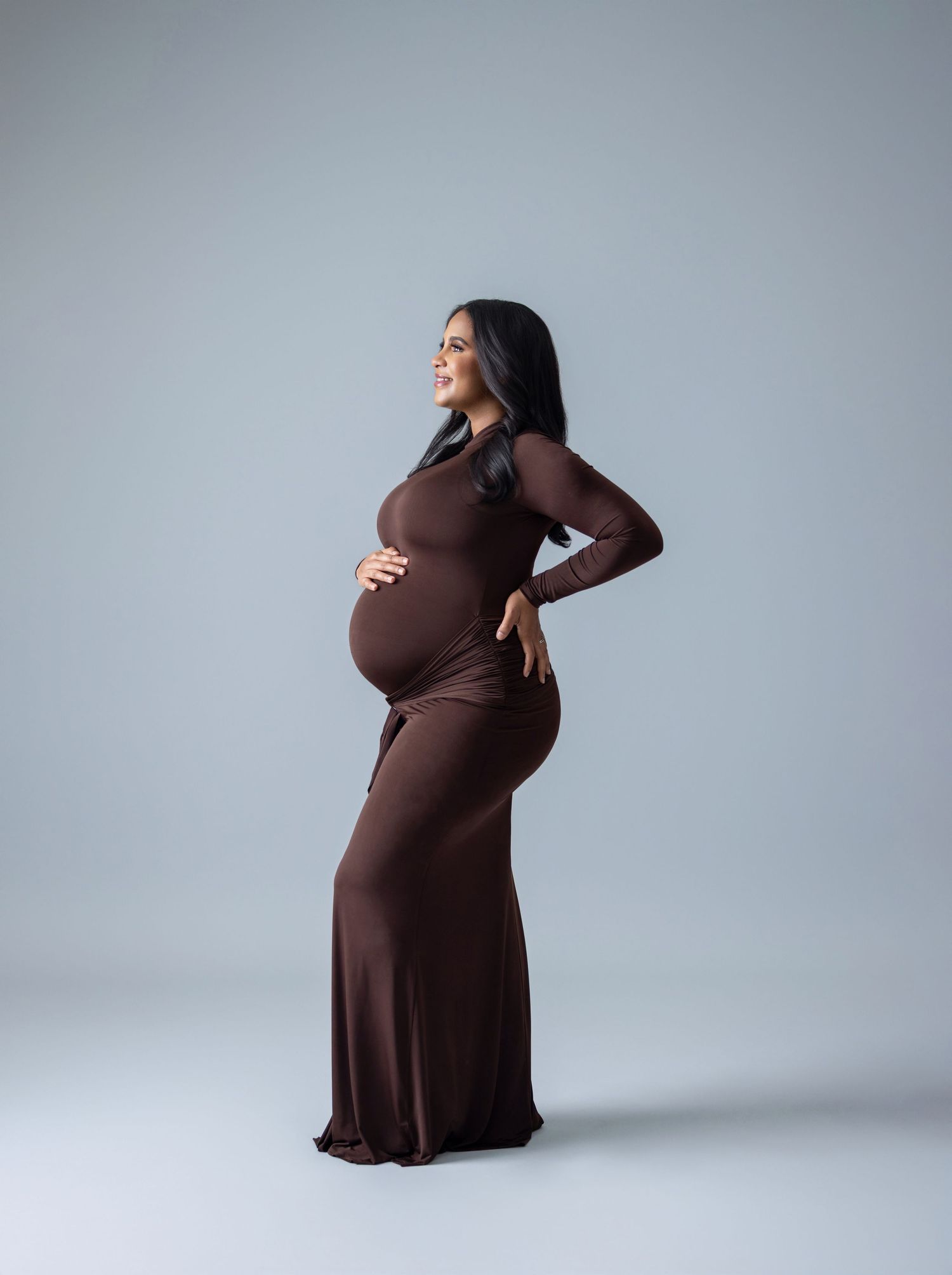 Pregnant woman in brown long-sleeve fitted maternity gown posing sideways, cradling baby bump in professional studio photo.
