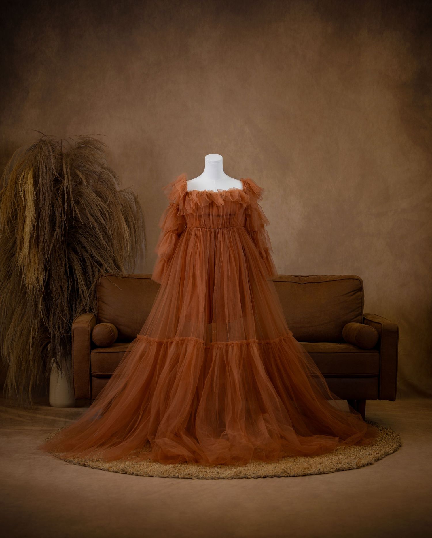 Elegant rust-colored tulle maternity gown on mannequin with flowing sleeves and tiered skirt in studio setting.