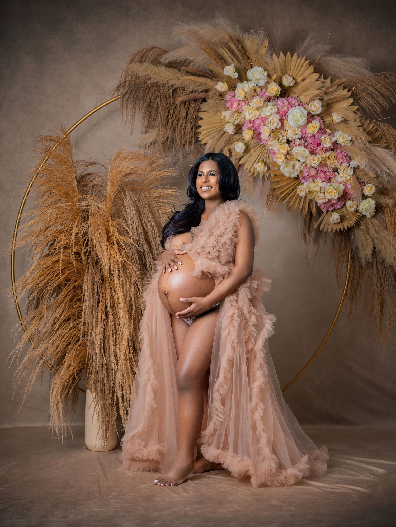 Pregnant woman in blush tulle robe poses beside floral hoop and pampas grass for elegant maternity photo.