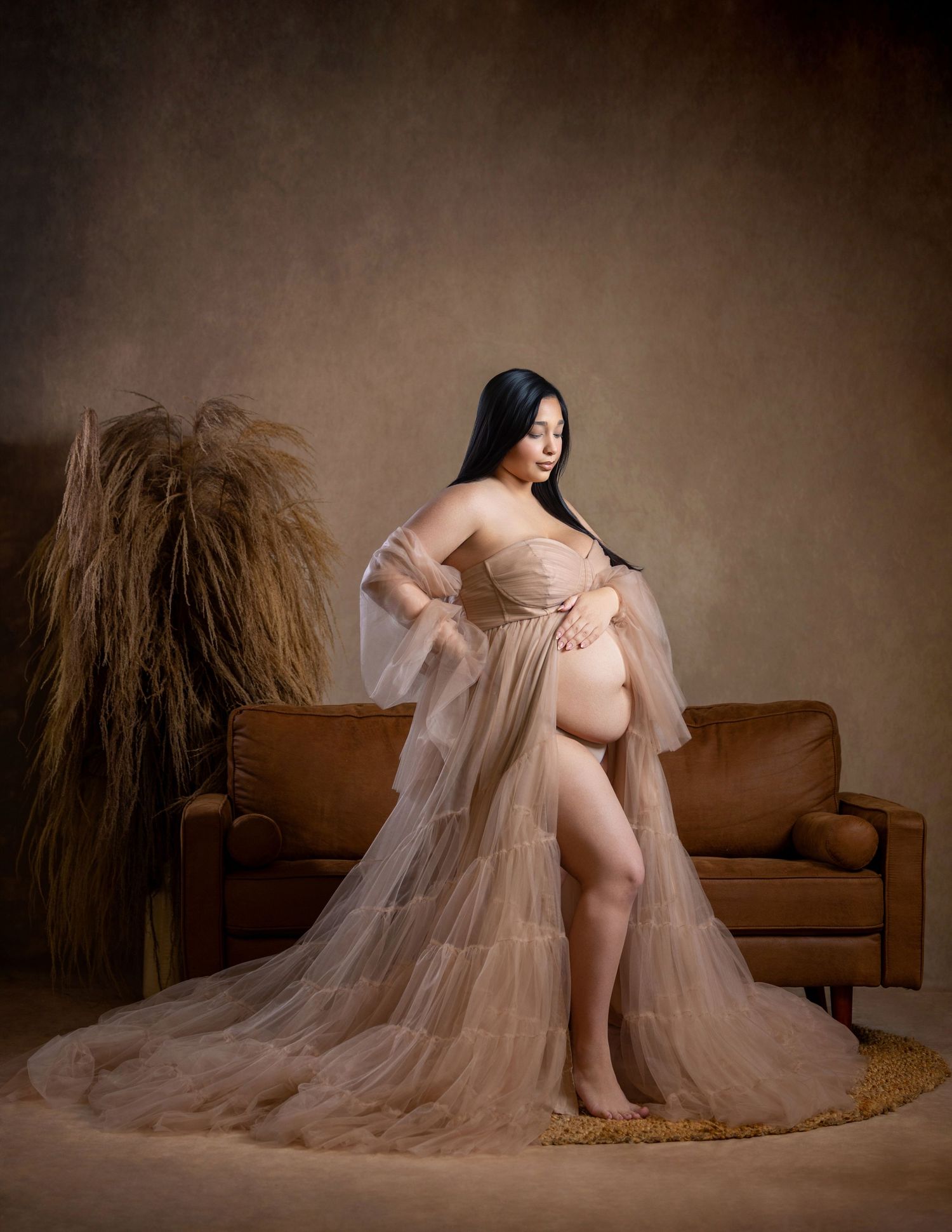 Pregnant woman in sheer beige tulle maternity gown posing elegantly in a warmly lit studio with leather sofa.