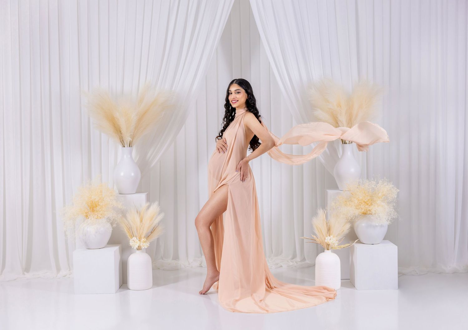 Woman in elegant peach flowing gown poses gracefully in white studio with pampas grass decor.