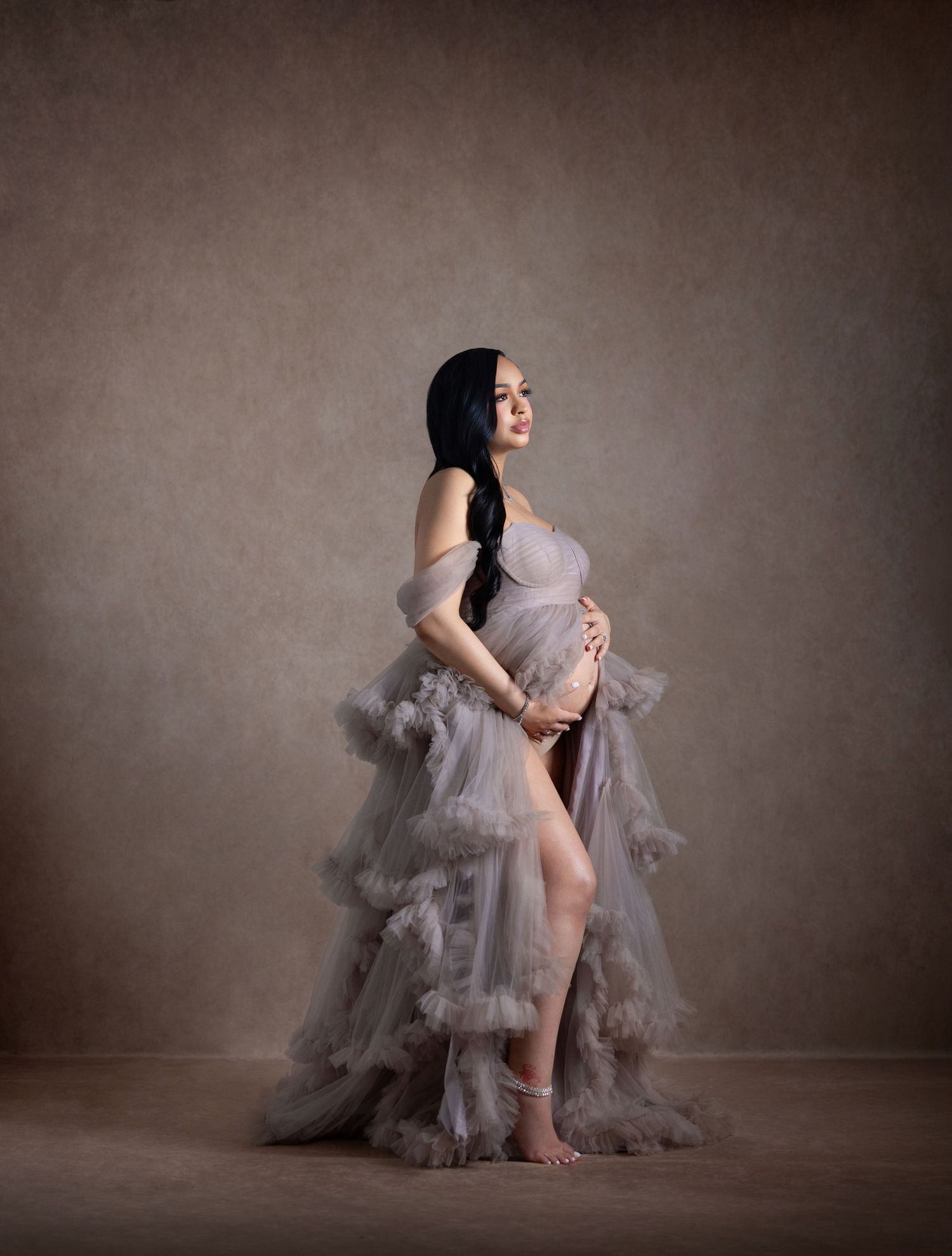 Pregnant woman in flowing gray tulle gown cradling baby bump in elegant studio maternity portrait.