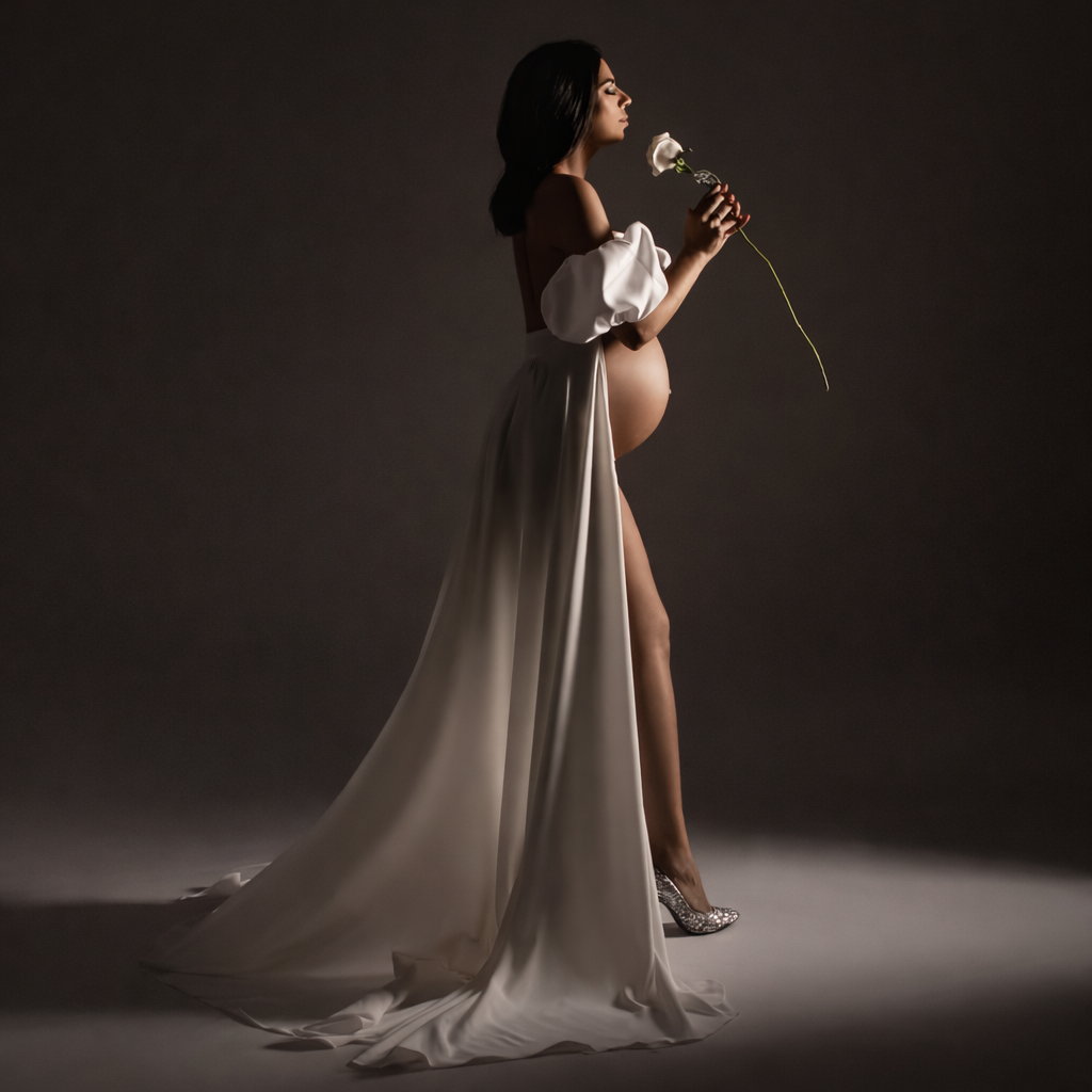 Pregnant woman in elegant white off-shoulder gown holding a rose, posing gracefully in a studio maternity portrait.