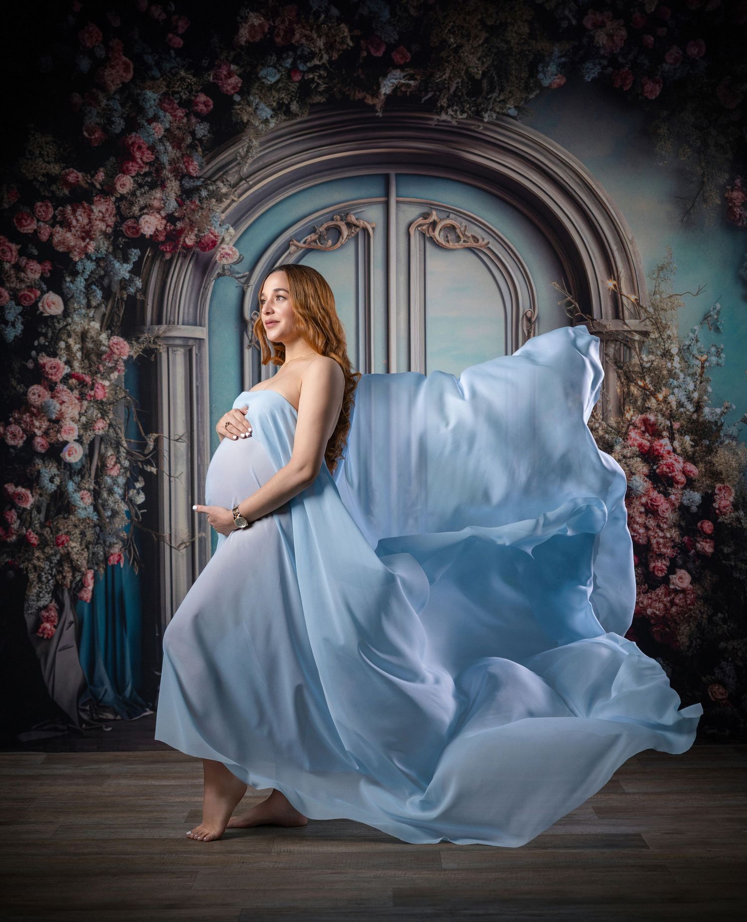 Pregnant woman in flowing light blue gown poses gracefully before a floral arch backdrop in a maternity photo session.
