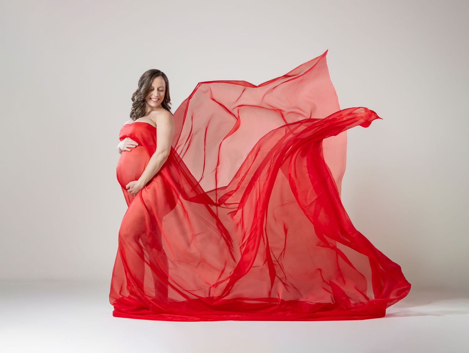 Pregnant woman in flowing red chiffon gown with fabric billowing dramatically against white studio background.