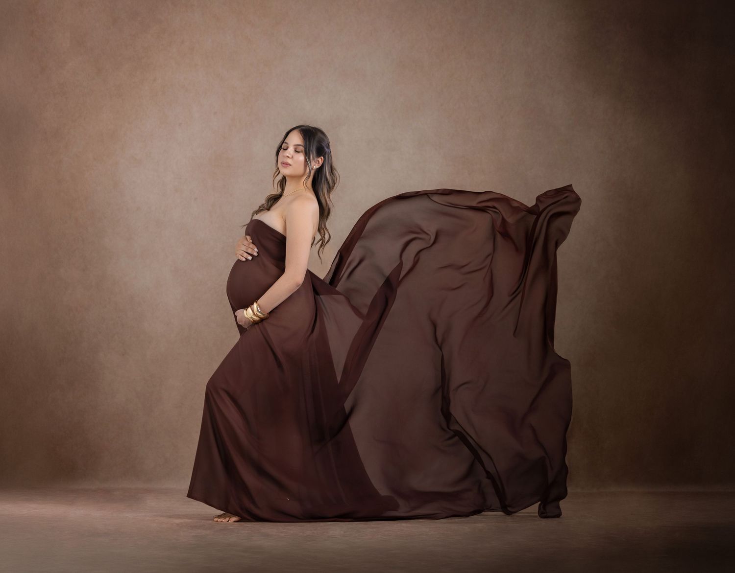 Pregnant woman in flowing brown maternity gown poses gracefully against a warm studio backdrop.