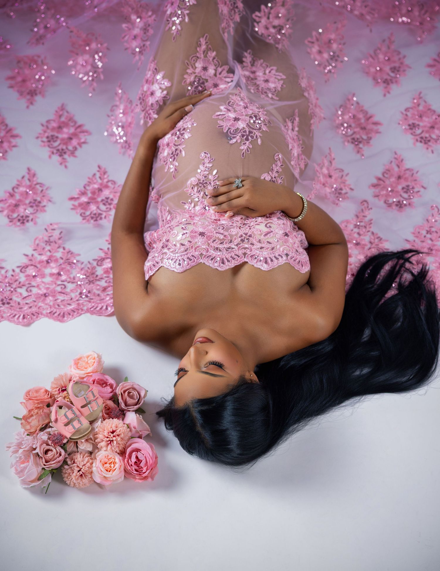 Pregnant woman in pink lace top lying on floor with roses and pink embroidered fabric backdrop.