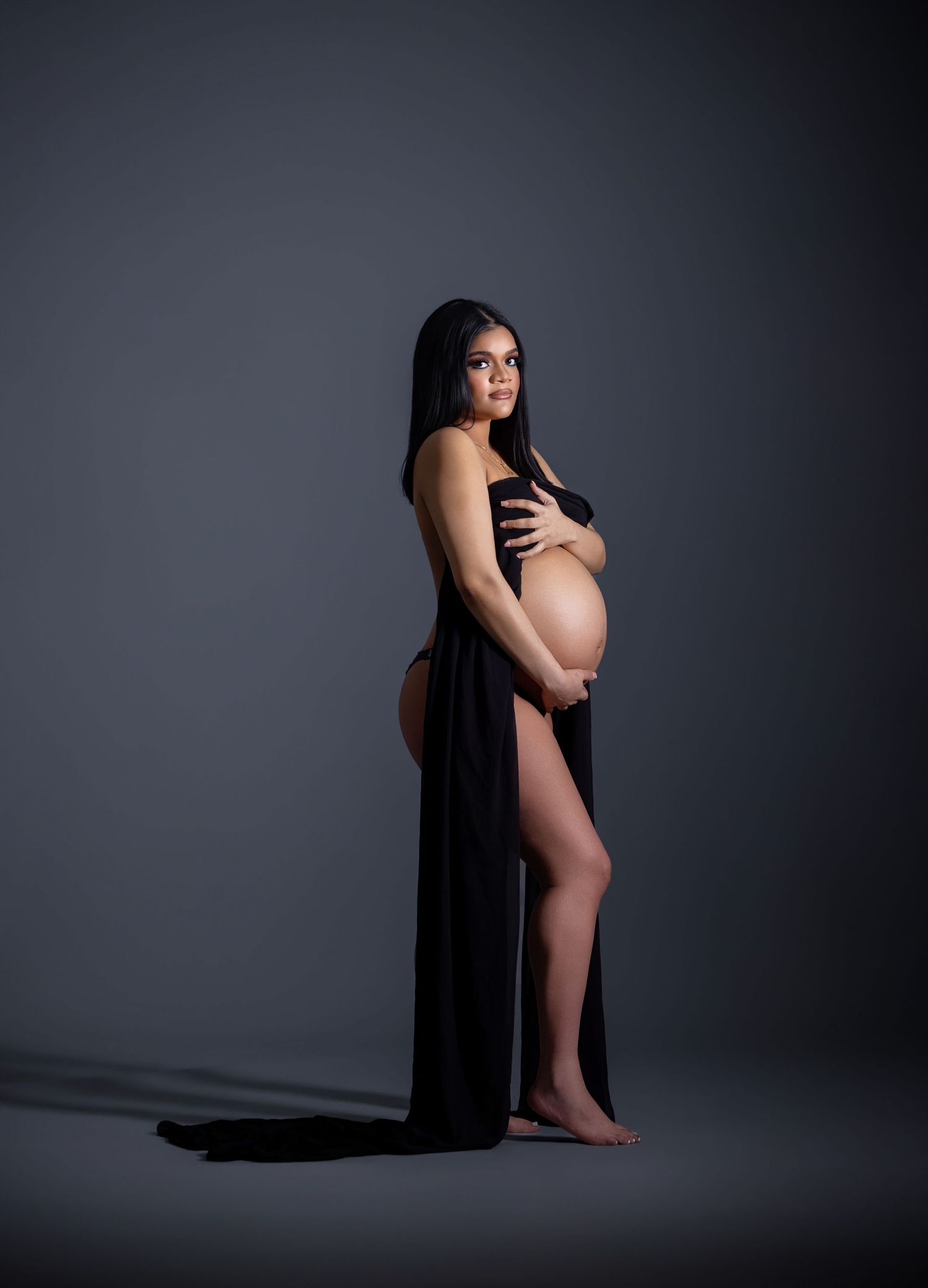 Pregnant woman in black draped gown posing sideways, cradling baby bump in professional maternity studio photo.