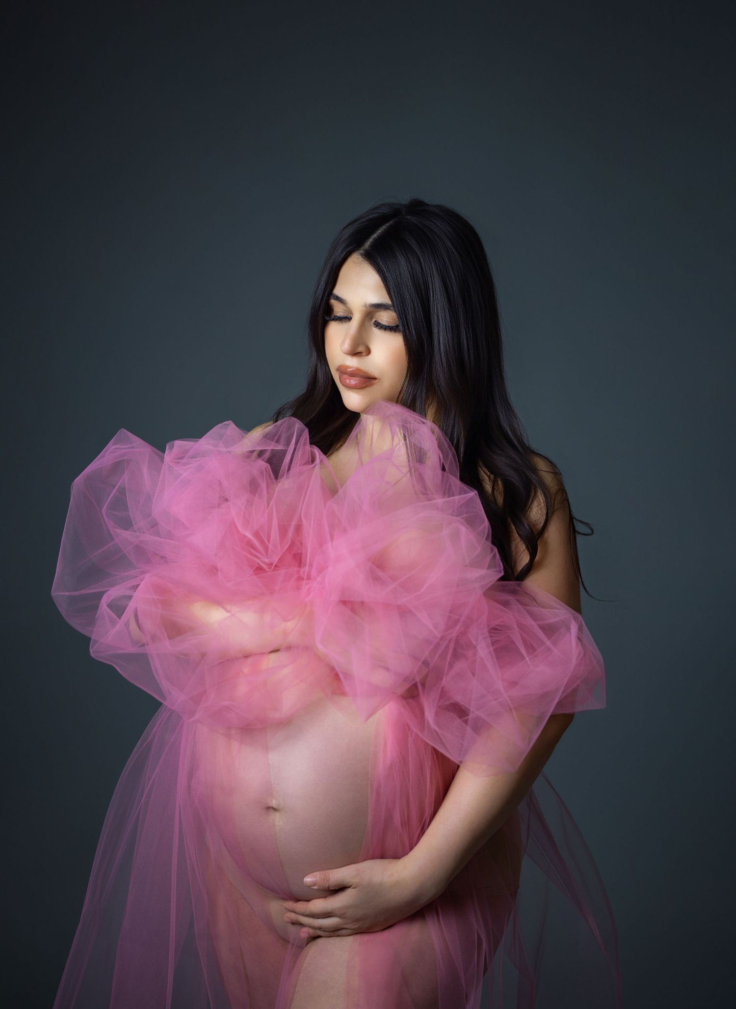 Pregnant woman in pink tulle robe cradling baby bump in professional maternity studio portrait.