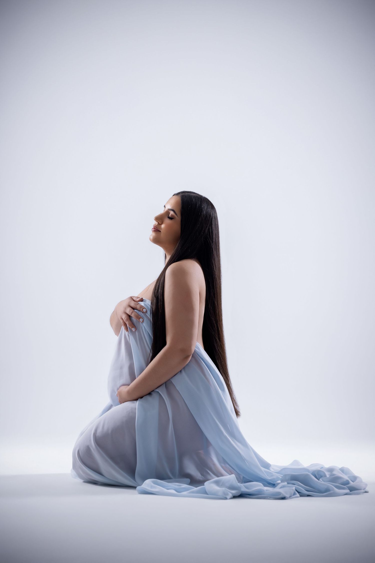 Pregnant woman kneeling in flowing blue sheer fabric draped elegantly, long dark hair, serene pose on white background.
