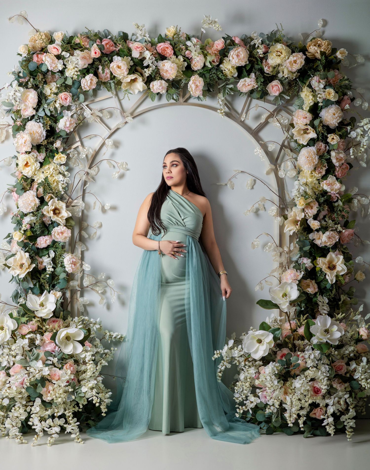 Pregnant woman in sage green tulle gown poses under floral arch with pink and white blooms in studio.