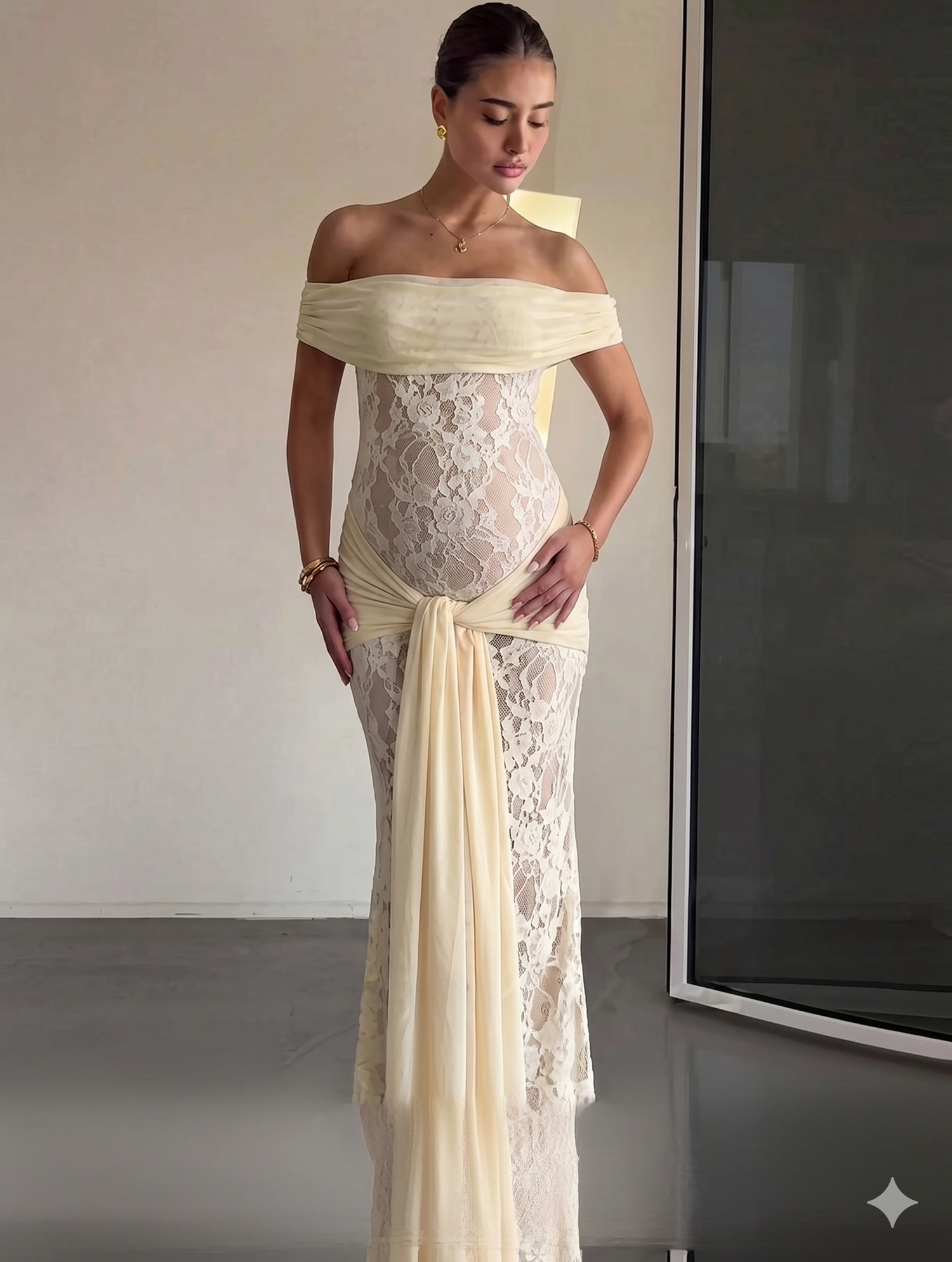 Pregnant woman in elegant off-shoulder cream lace maxi dress with chiffon sash, posing gracefully.