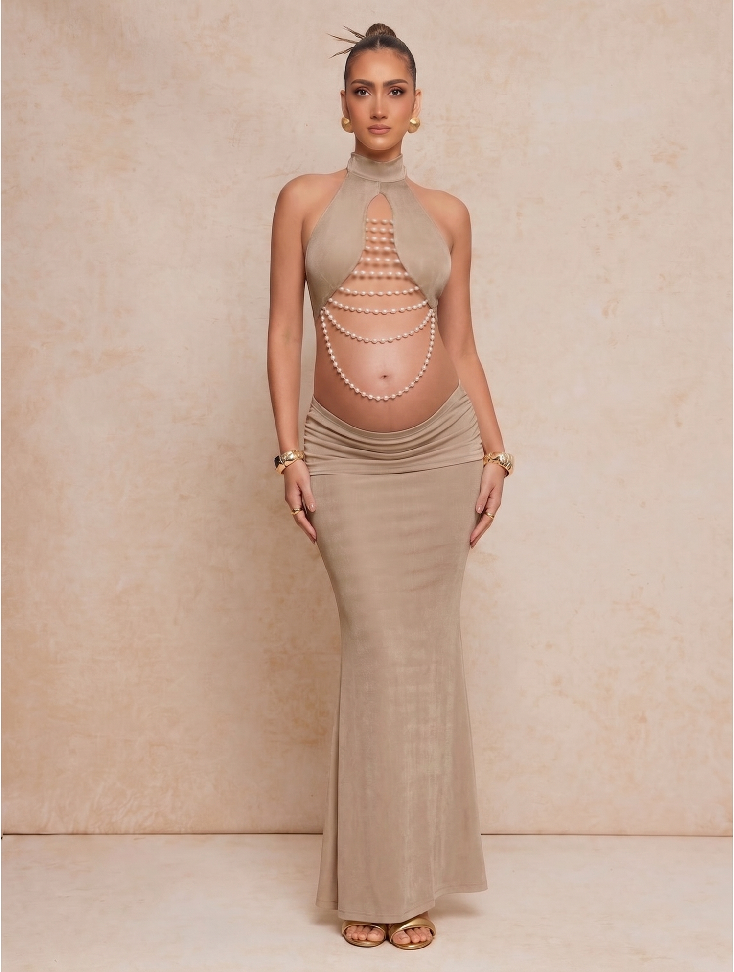 Pregnant woman in taupe halter maxi dress with pearl belly chain cutout detail, styled with gold accessories.