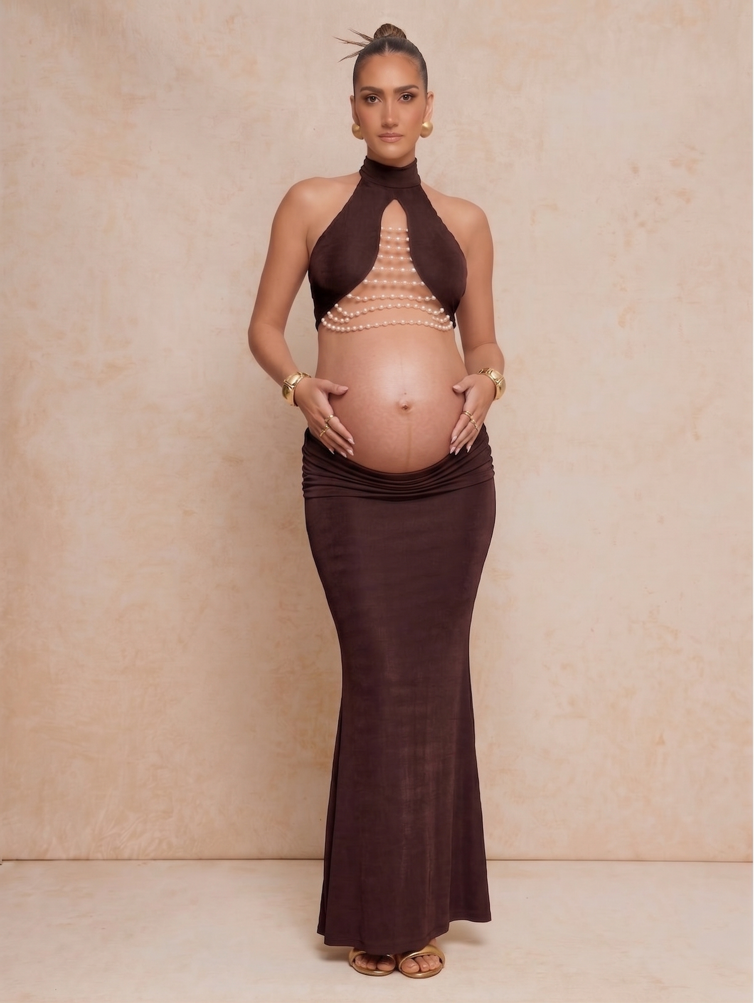 Pregnant woman in brown halter maxi dress with pearl embellishments, showcasing baby bump in elegant maternity fashion.