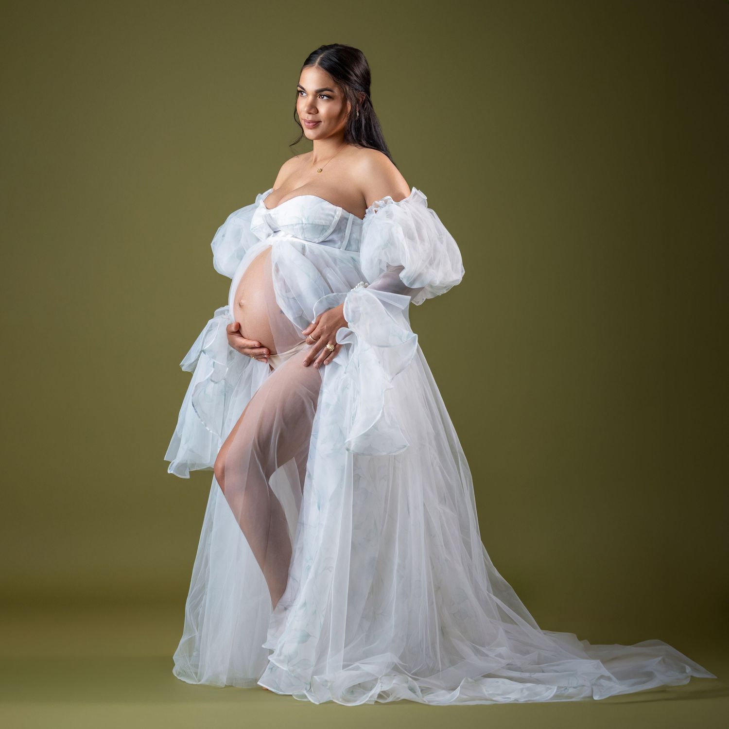 Pregnant woman in elegant white off-shoulder tulle maternity gown with puff sleeves posing against olive background.