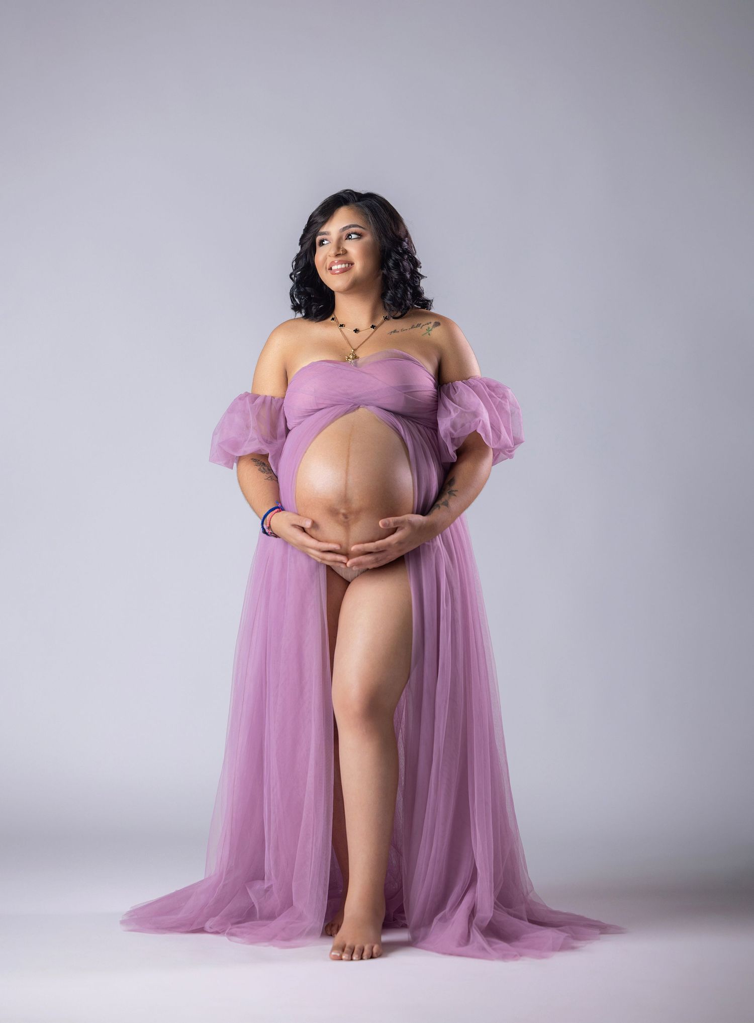 Pregnant woman in elegant lavender tulle maternity gown cradling baby bump in professional studio photo.