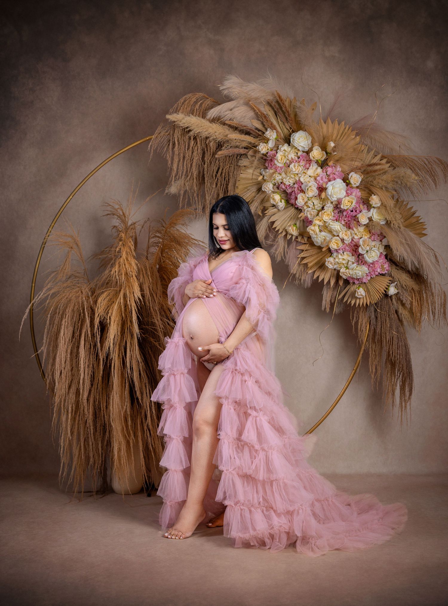 Pregnant woman in pink ruffle maternity gown poses before floral hoop arch with dried pampas grass.