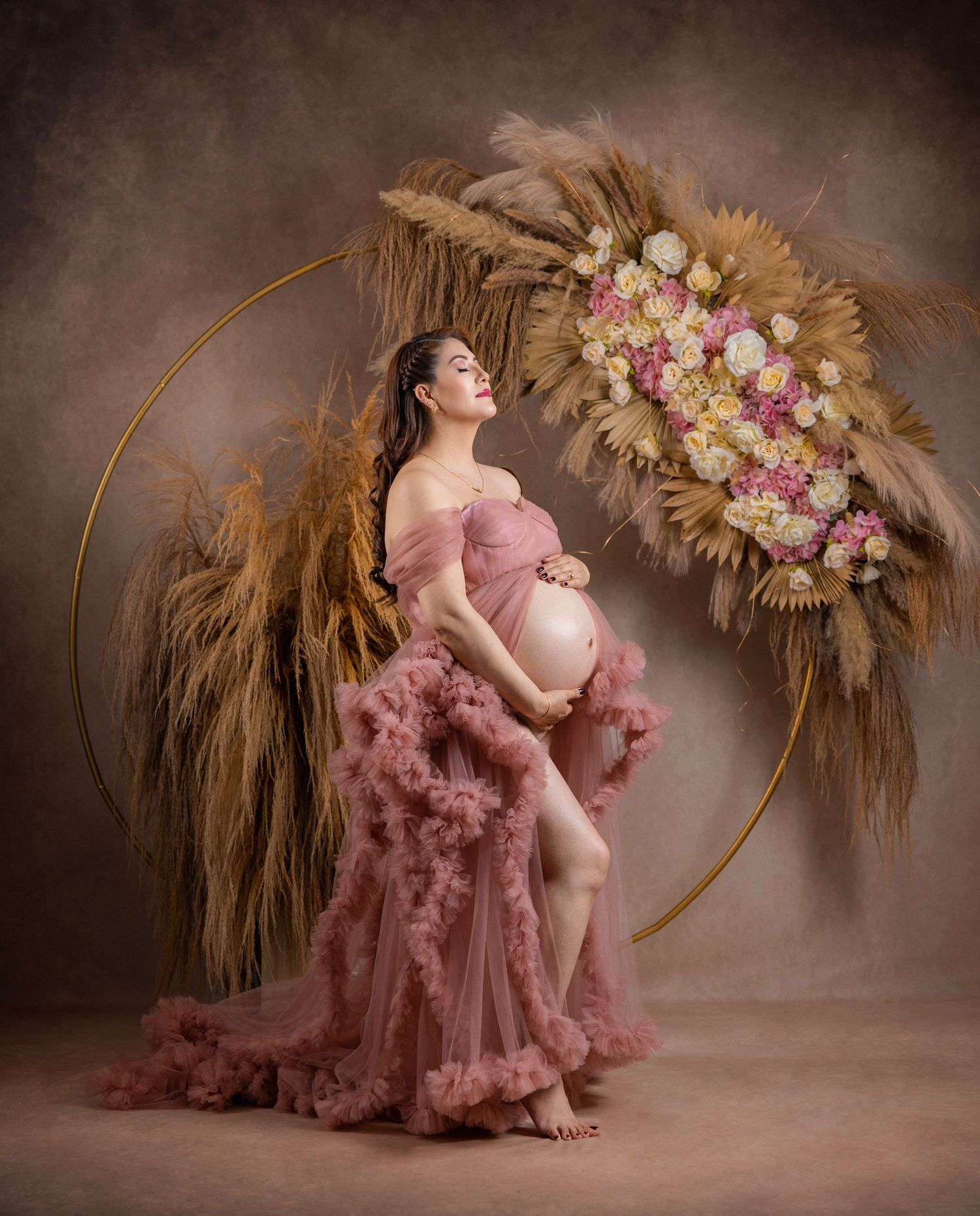 Pregnant woman in pink tulle gown poses gracefully before a floral hoop with pampas grass in a studio.