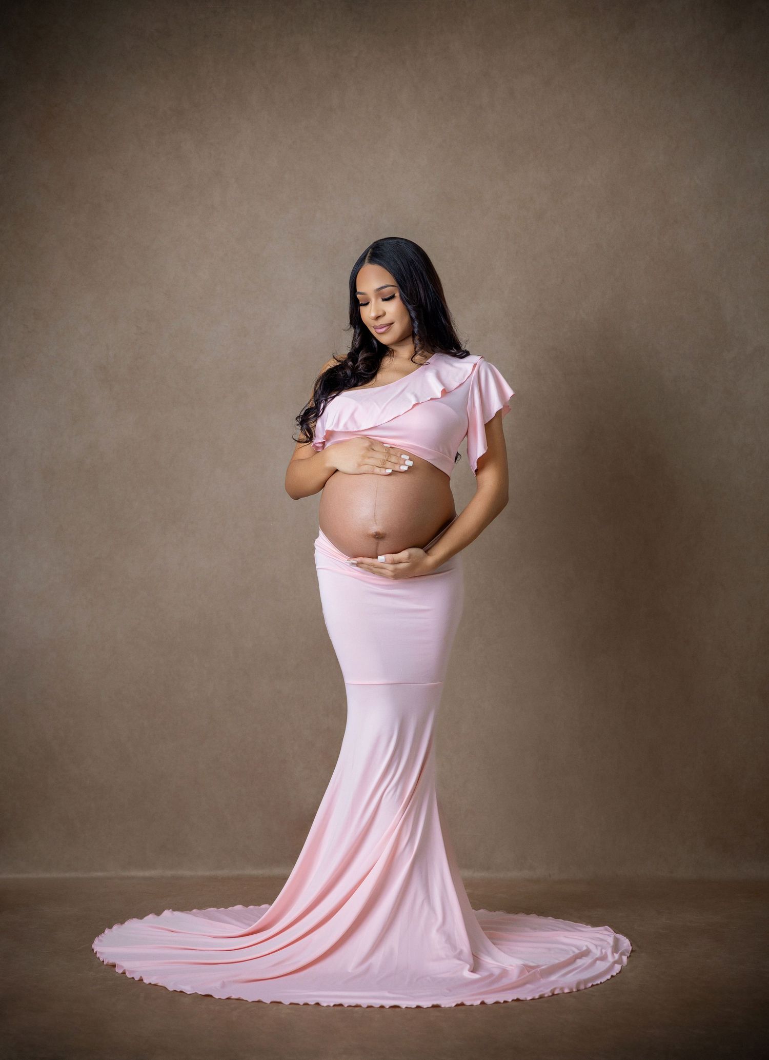Pregnant woman in pink off-shoulder maternity gown cradling baby bump in elegant studio portrait.