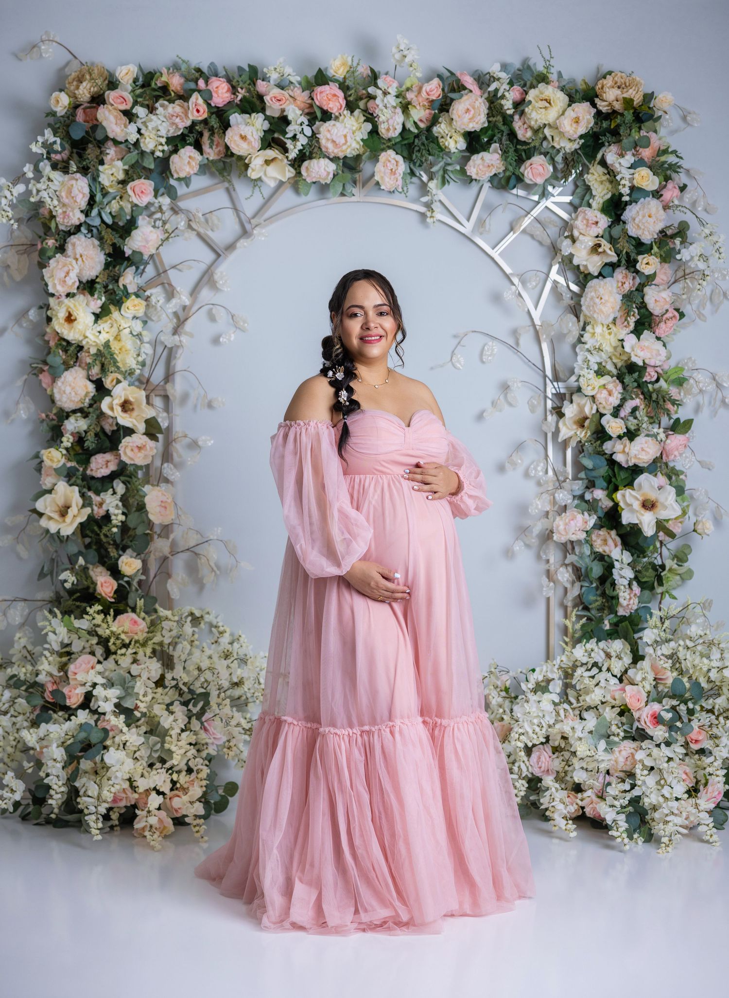 Pregnant woman in pink off-shoulder tulle gown poses before floral arch with roses for maternity photo.