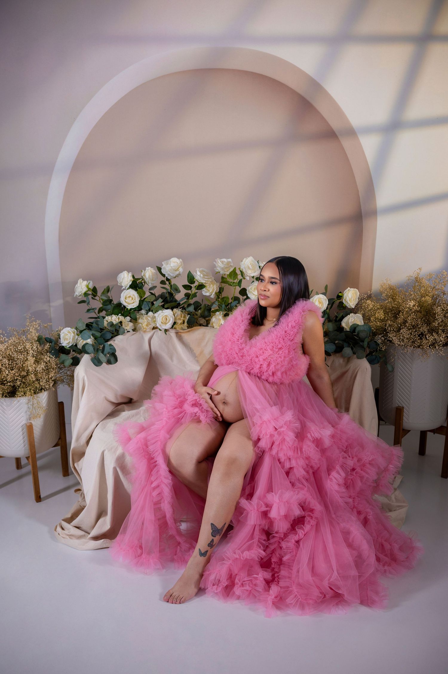 A woman in a fluffy pink tulle maternity gown poses gracefully in a studio with white roses and elegant decor.