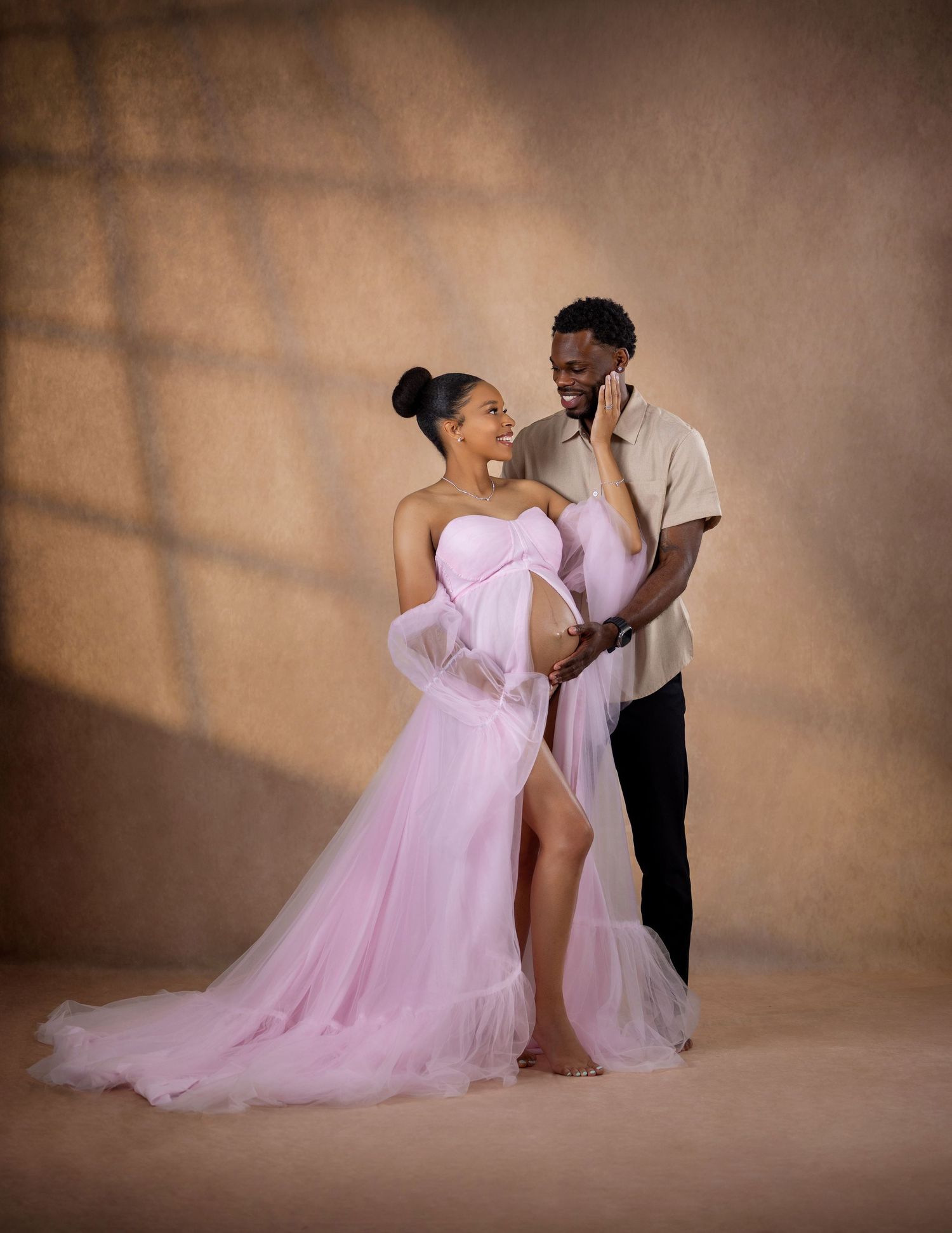 Pregnant woman in pink tulle maternity gown poses with partner in warm studio portrait setting.