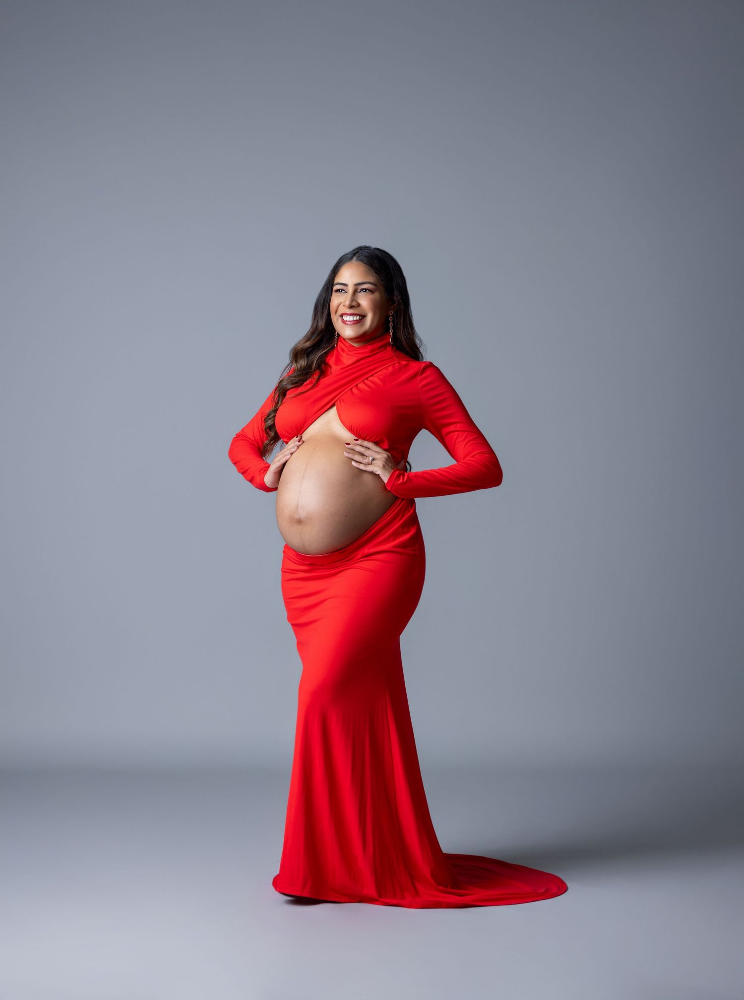 Smiling pregnant woman in a red cutout maternity gown posing in a studio for a maternity photoshoot.