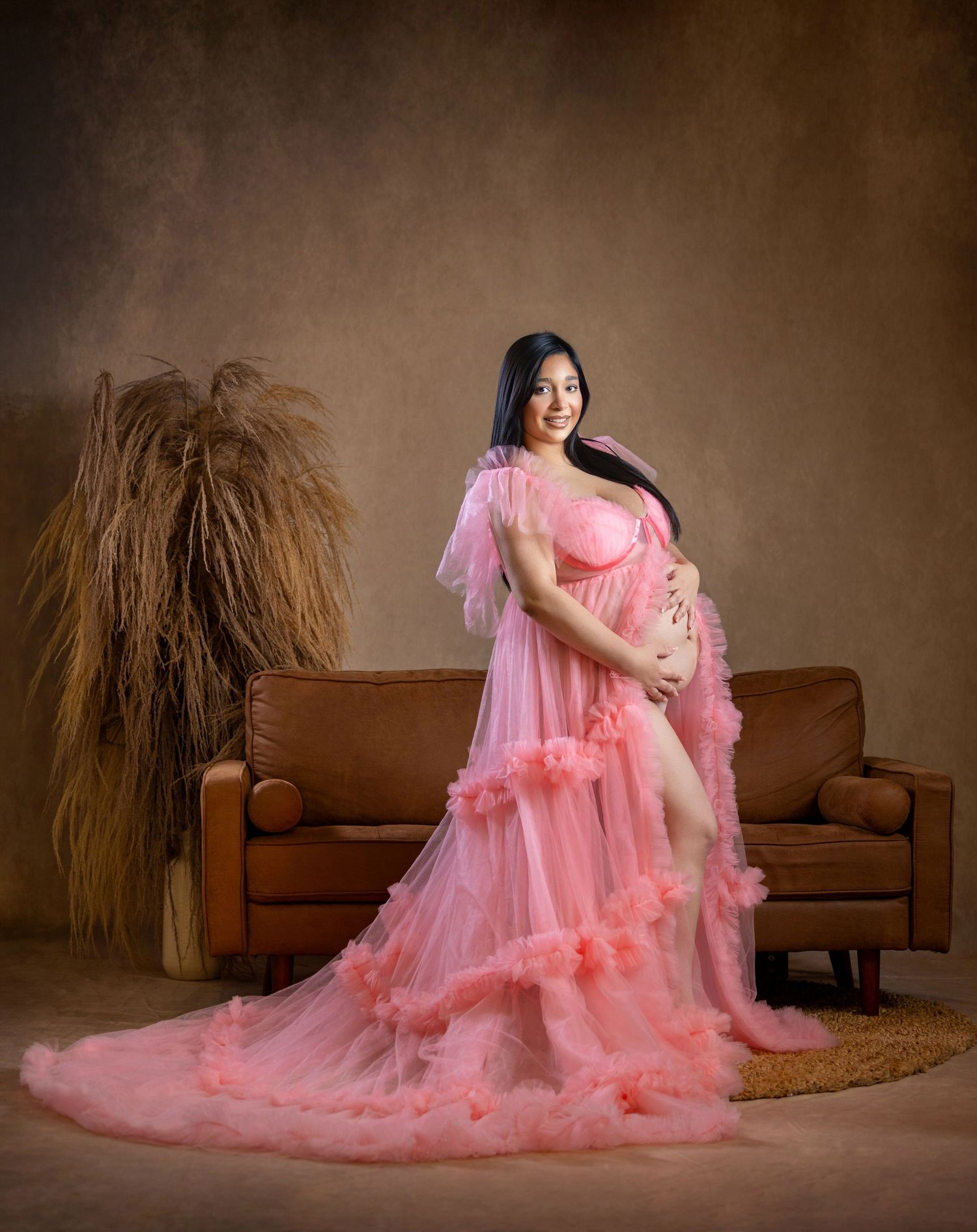 Pregnant woman in flowing pink tulle maternity gown posing elegantly in a studio with warm brown tones.