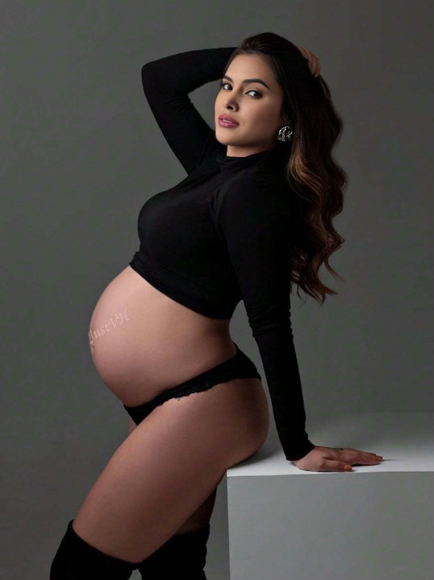 Pregnant woman in black crop top and thigh-high boots posing for a maternity photo shoot against gray background.