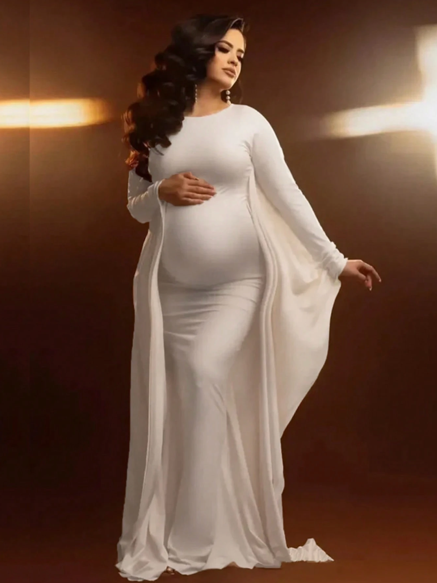 Pregnant woman in elegant white long-sleeve maxi dress with flowing cape sleeves, posing against warm brown background.