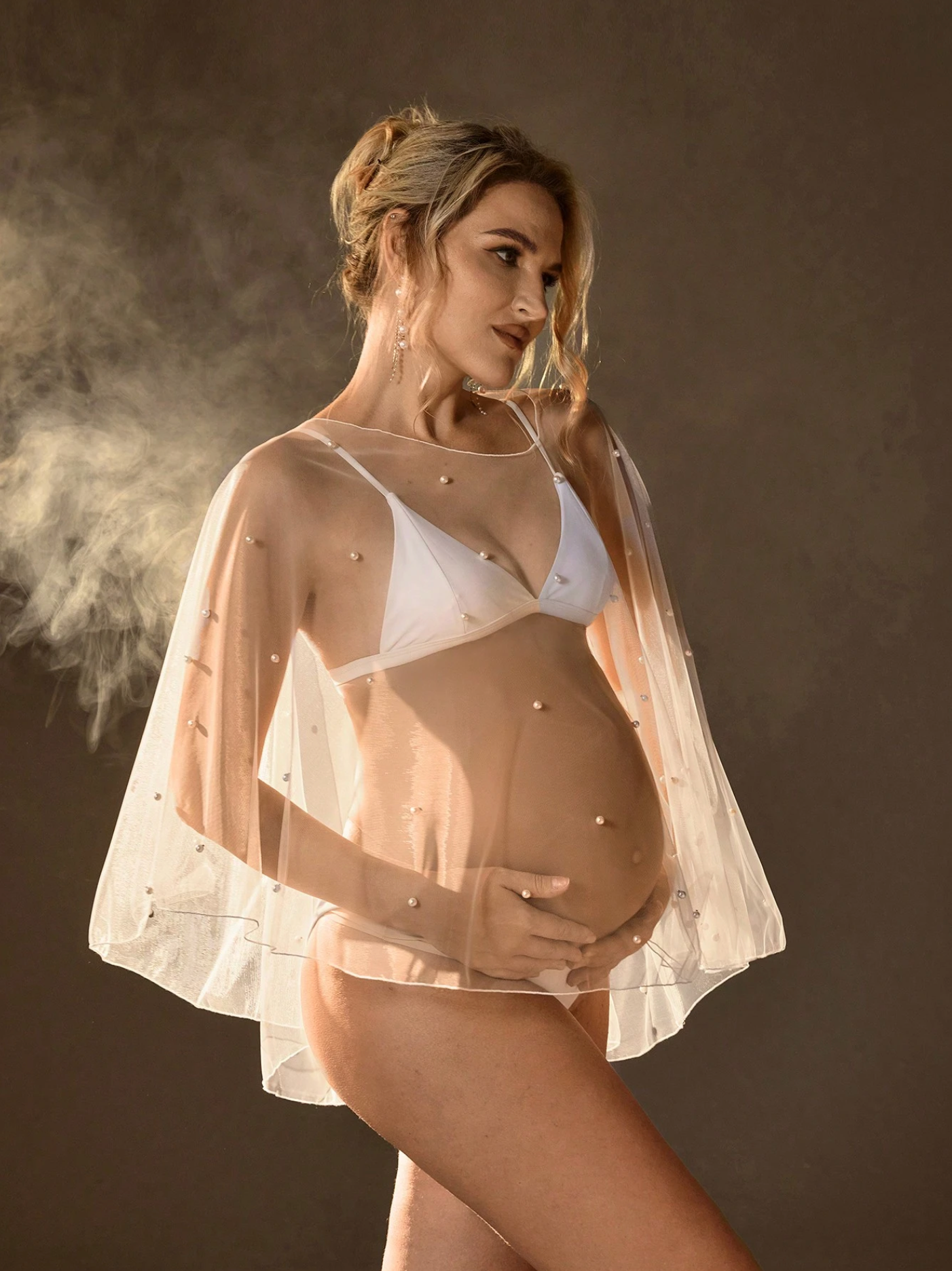 Pregnant woman in white bikini and sheer embellished cape poses gracefully in a moody studio setting.