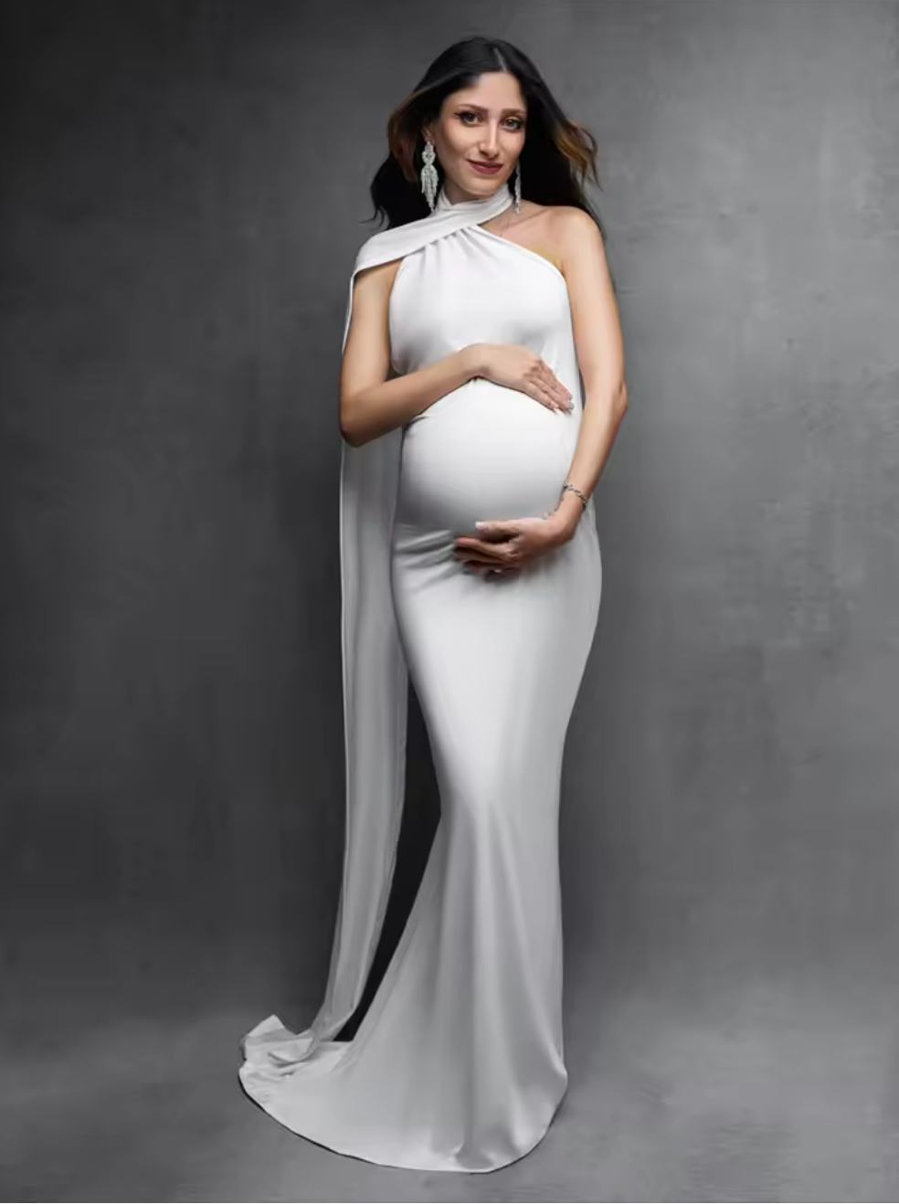 Pregnant woman in elegant white halter neck maternity gown with cape detail, posing gracefully against gray background.