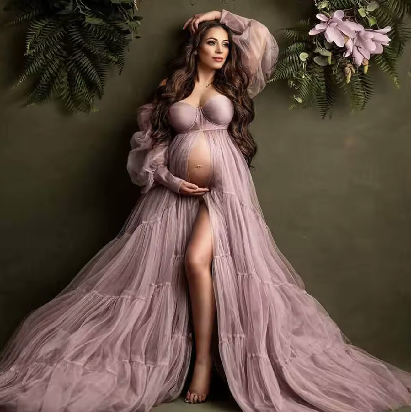 Pregnant woman in elegant dusty pink tulle maternity gown with puff sleeves, posing gracefully against floral backdrop.
