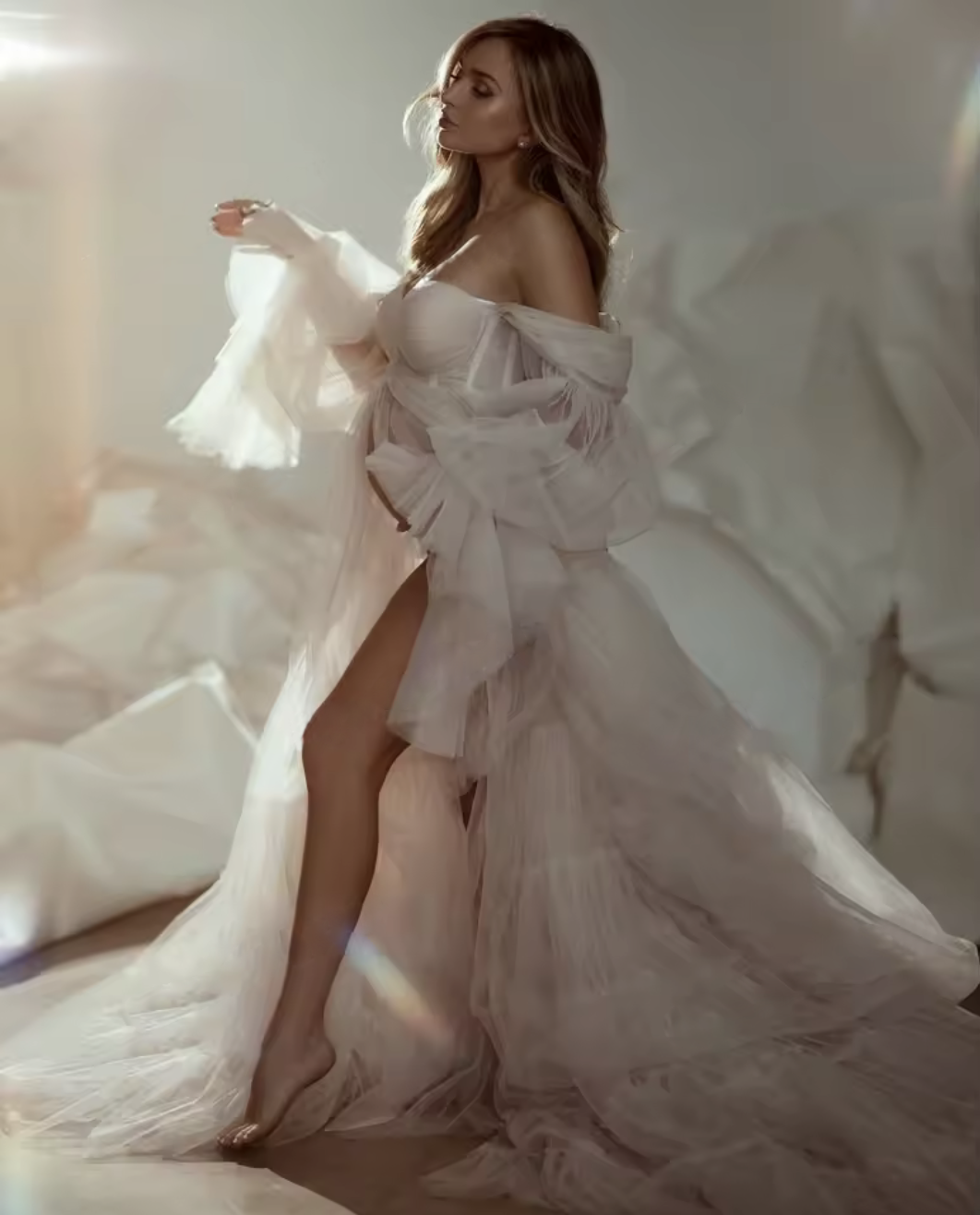 Woman in flowing off-shoulder white tulle gown poses elegantly in soft natural light.