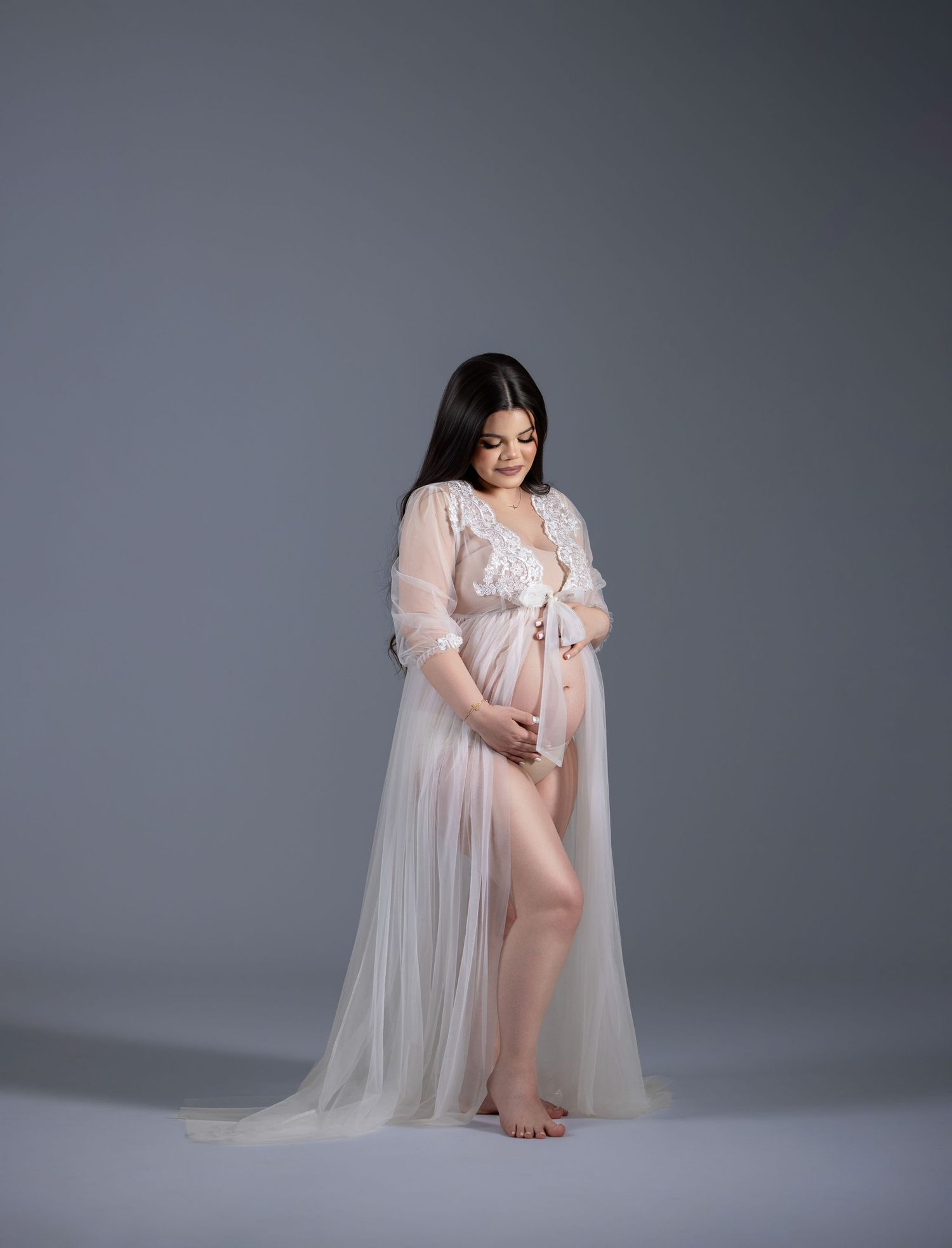 Pregnant woman in sheer white maternity gown cradling baby bump in professional studio photography session.