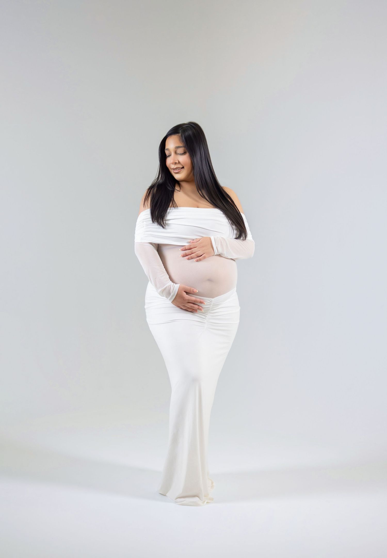 Pregnant woman in white off-shoulder maternity gown cradling her baby bump during a professional maternity photoshoot.