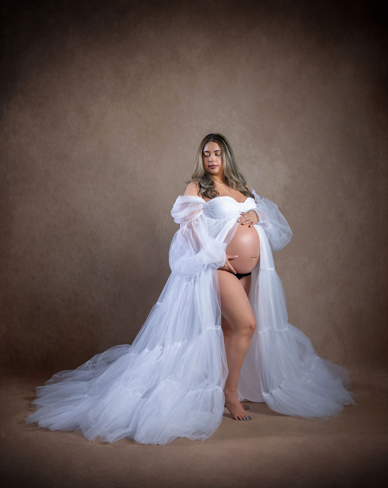 Pregnant woman in elegant white tulle maternity gown poses gracefully in professional studio photography session.