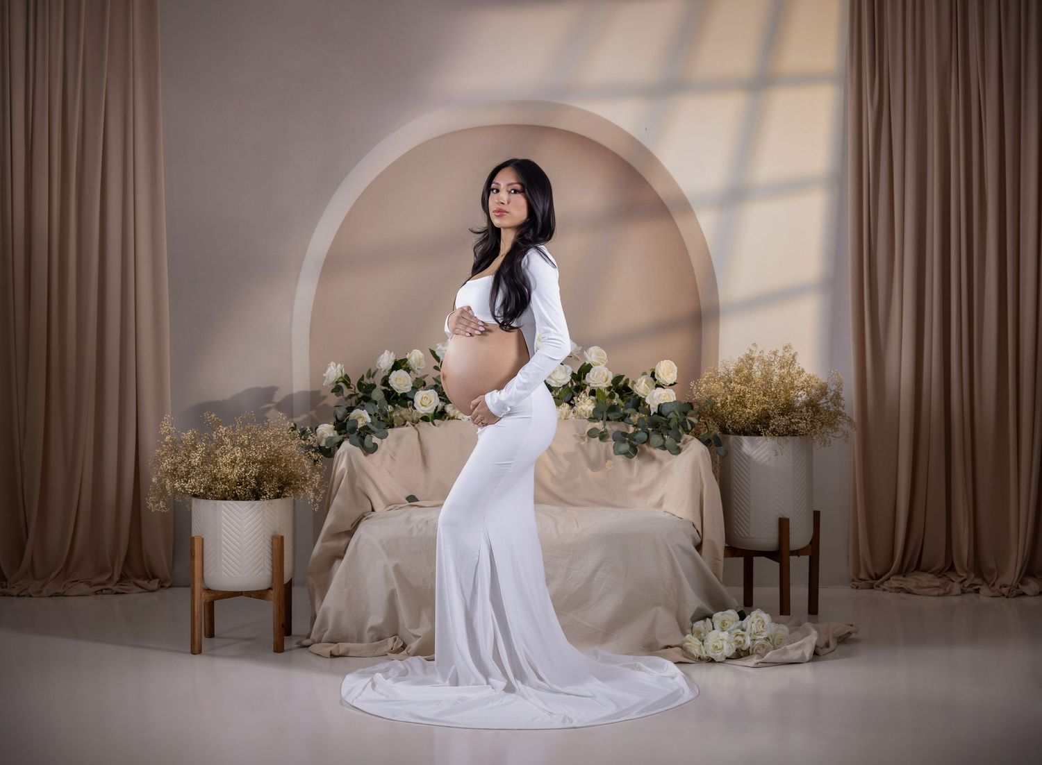 Pregnant woman in white gown posing in elegant studio with floral arrangements and arch backdrop.